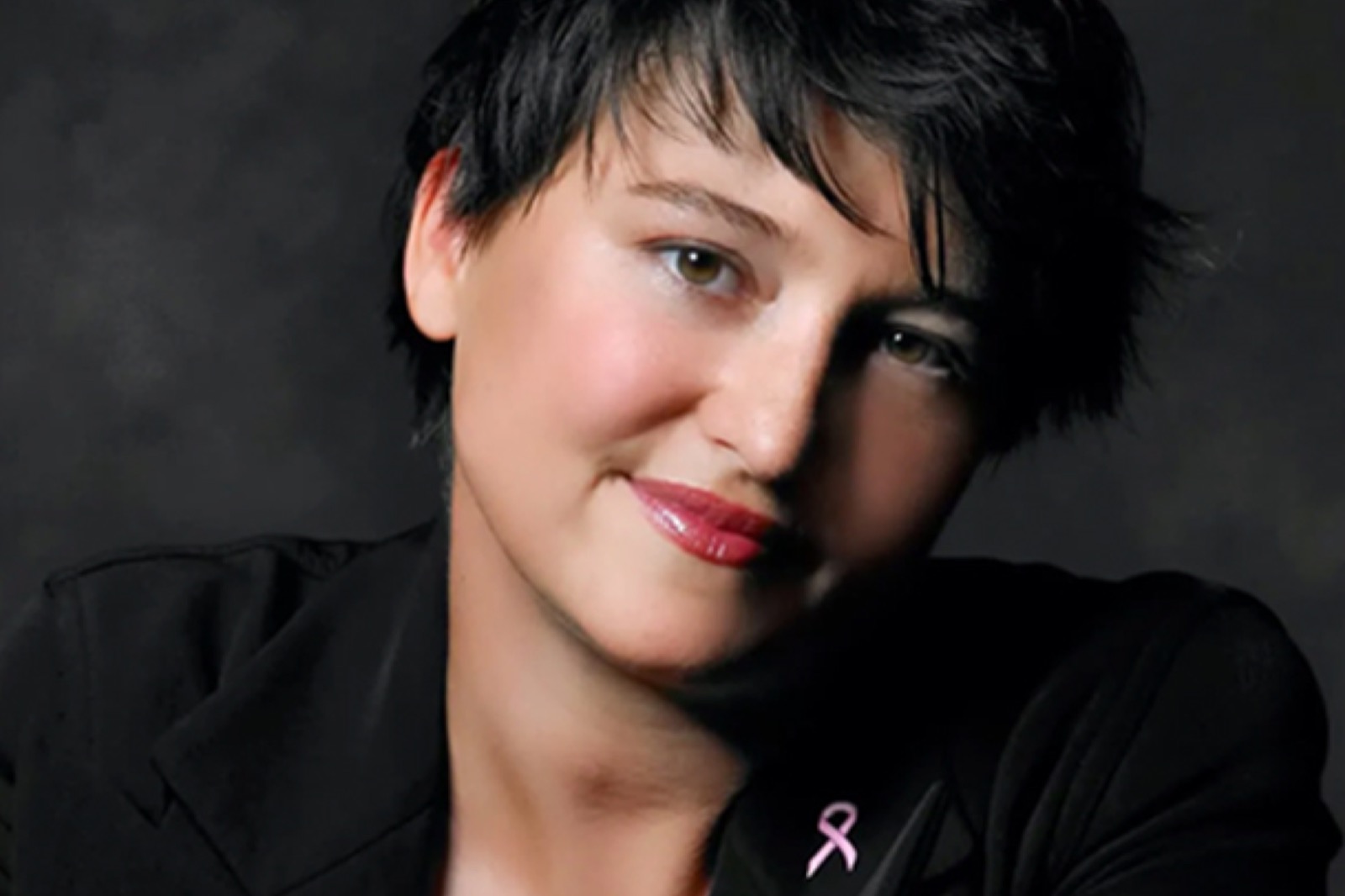 Woman slightly smiling at camera wearing a pink breast cancer awareness pin on collar.